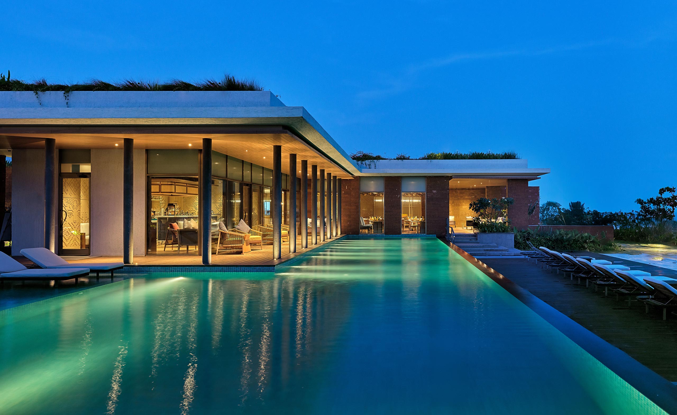 Modern villa at dusk with large glass windows and an illuminated interior, surrounded by a serene, expansive pool reflecting the deep blue sky.