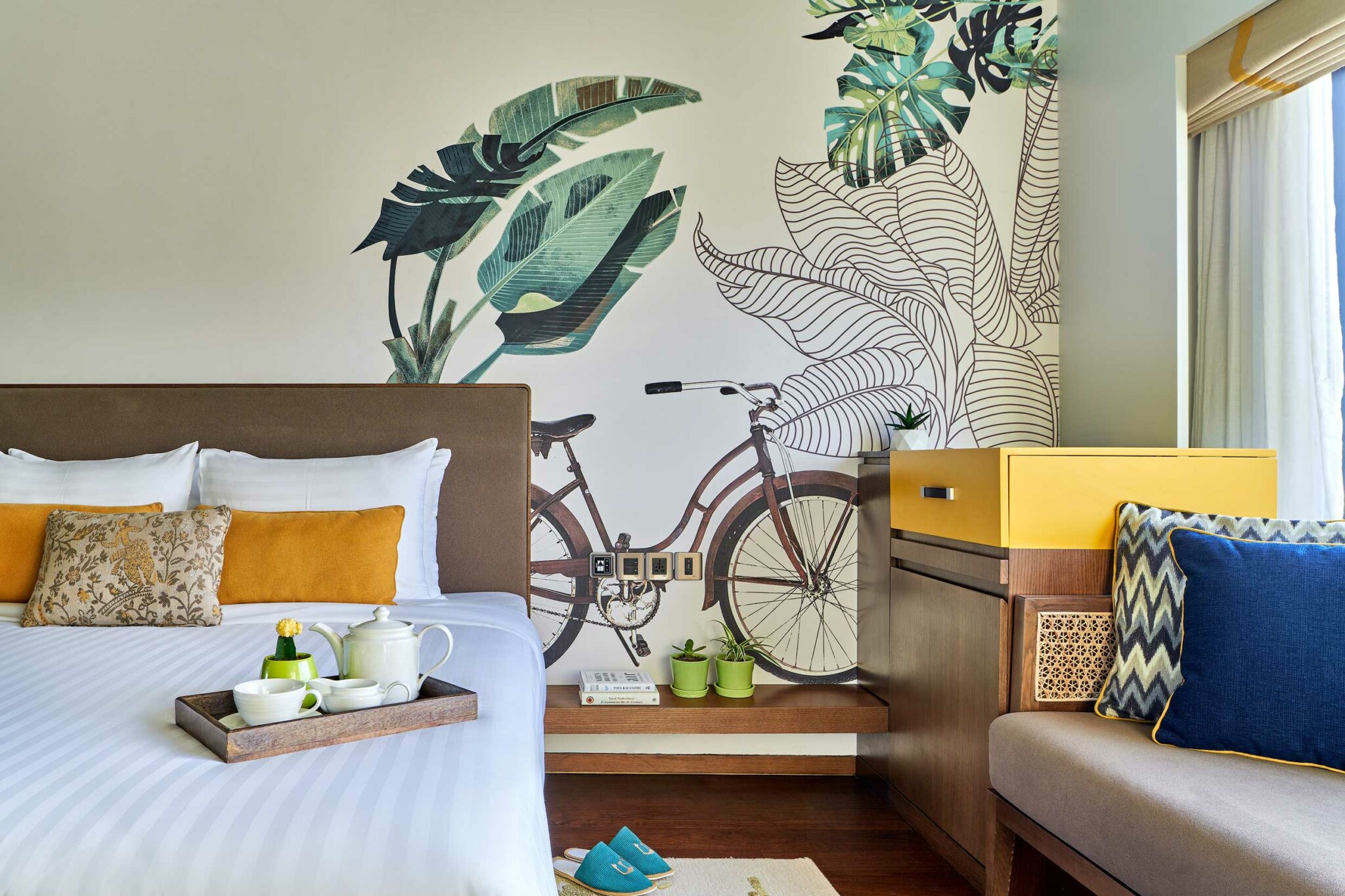 Bright room with a tropical theme features a bed with white and yellow pillows, bicycle wall art, and a wooden bench. A tray with a teapot rests on the bed.