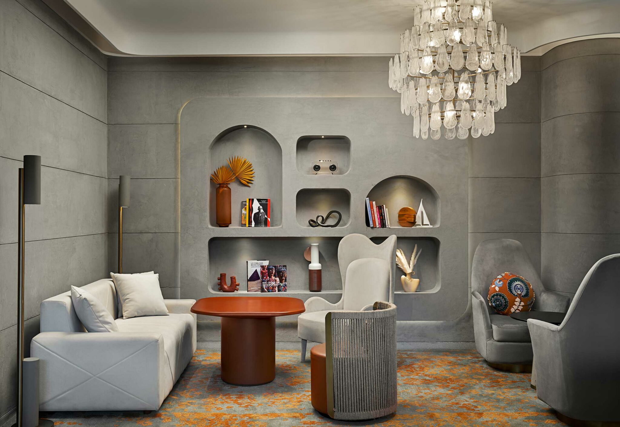 Sophisticated, modern lounge with gray curved walls and built-in shelves displaying art pieces. Features plush seating, vibrant rug, and elegant chandelier.