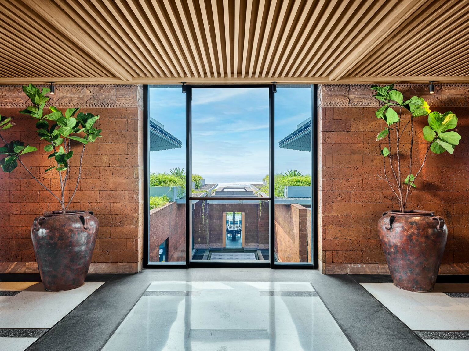 Modern interior with a wooden ceiling and checkered floor, featuring two large potted plants flanking a glass door. The door offers a serene ocean view.