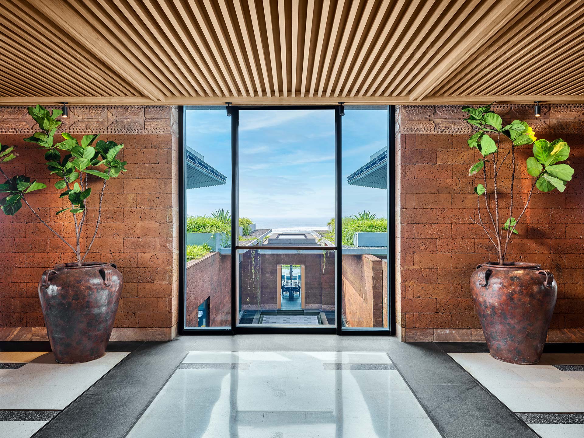 Modern interior with a wooden ceiling and checkered floor, featuring two large potted plants flanking a glass door. The door offers a serene ocean view.