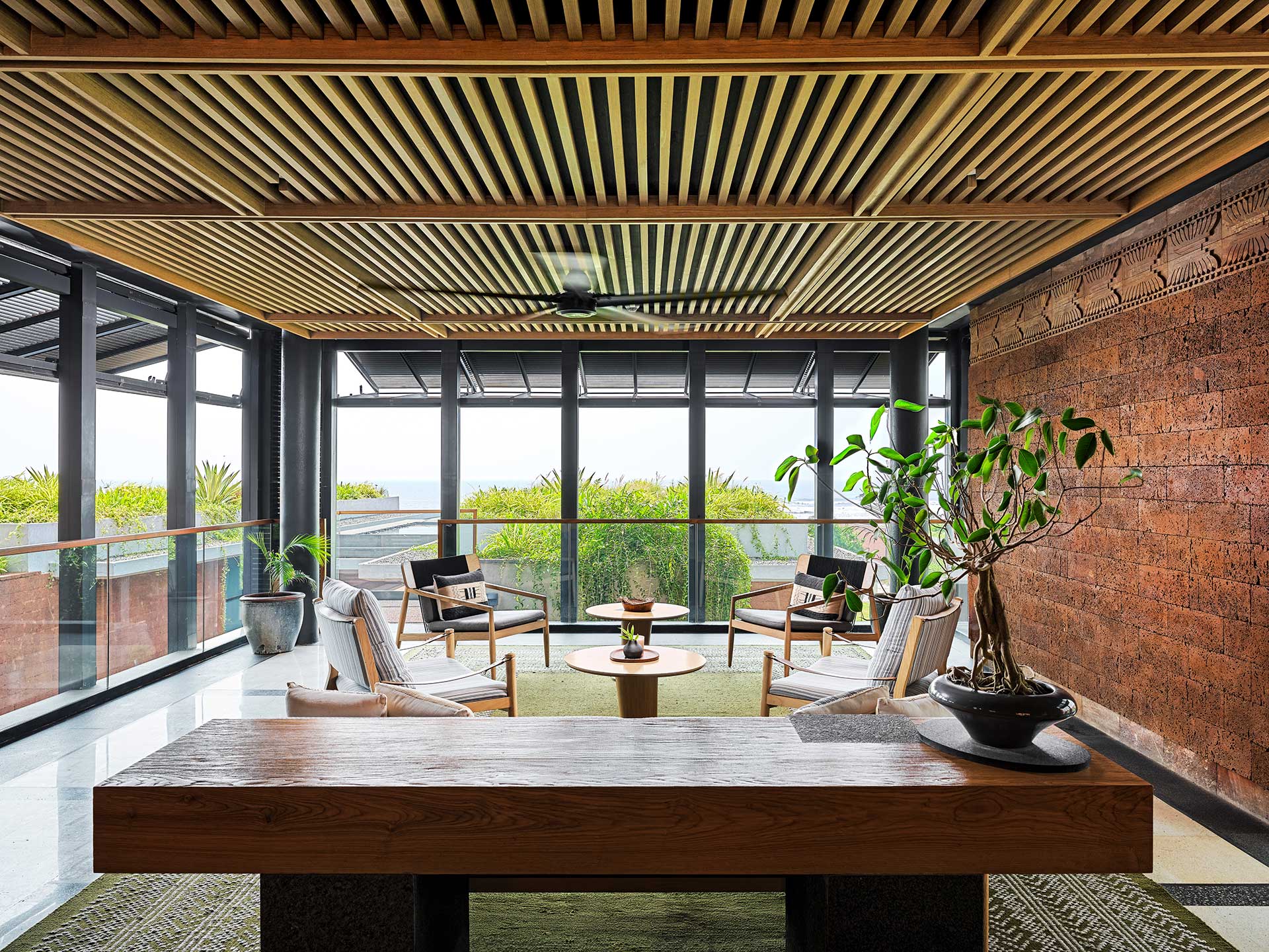 Modern interior with wooden slatted ceiling, large windows, and brick wall. Features potted plants, a wooden table, and four armchairs around a round table.