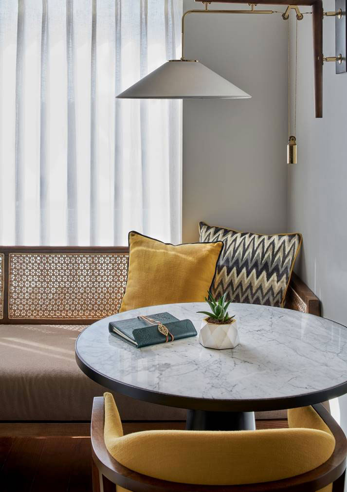 A cozy nook with a round marble table, flanked by a mustard chair. A bench has mustard and zigzag pillows, near sheer curtains and a sleek wall lamp.