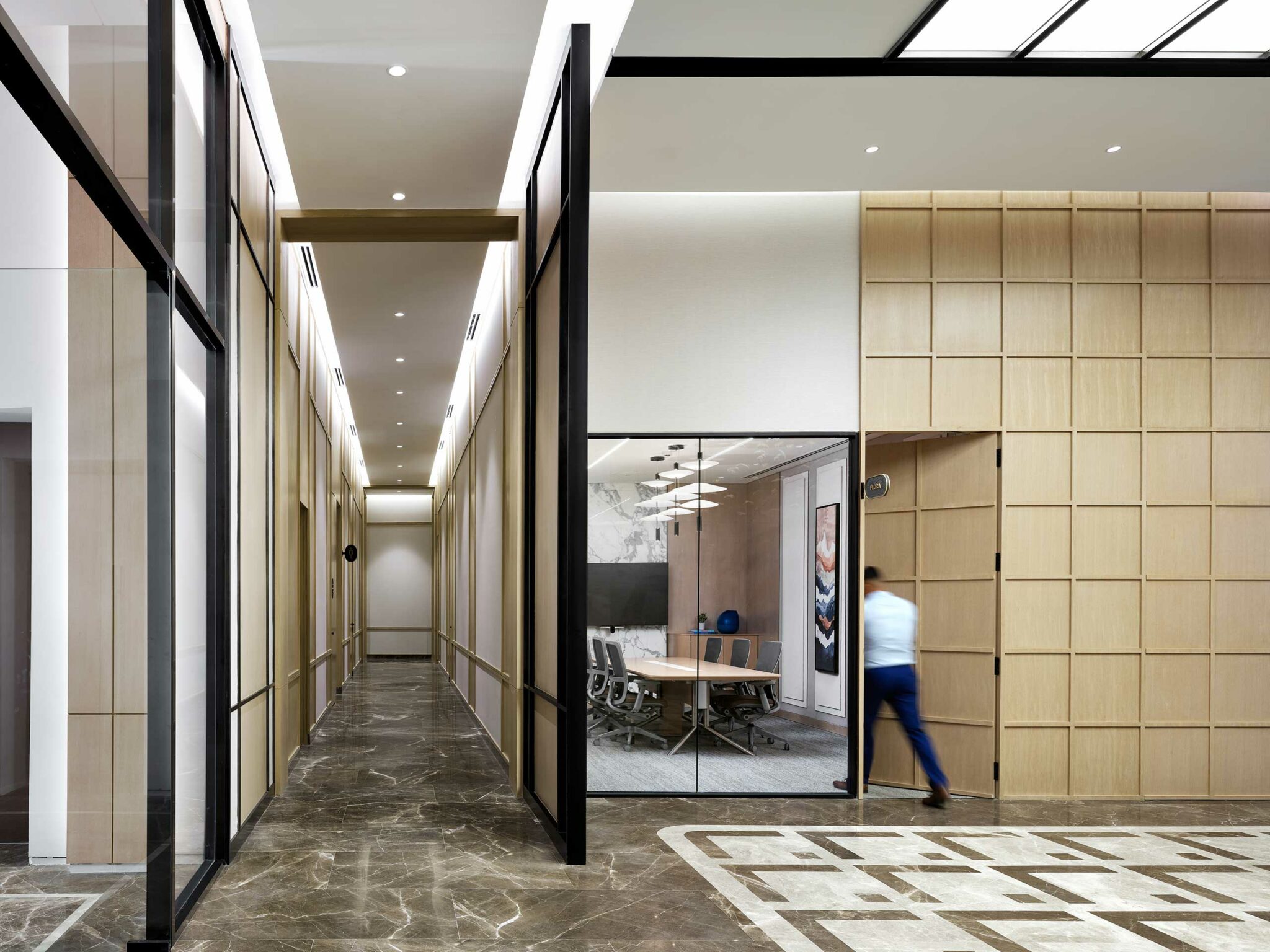 Modern office hallway with glass walls and a marble floor. A meeting room with a conference table is visible through a glass door. A person is entering on the right.