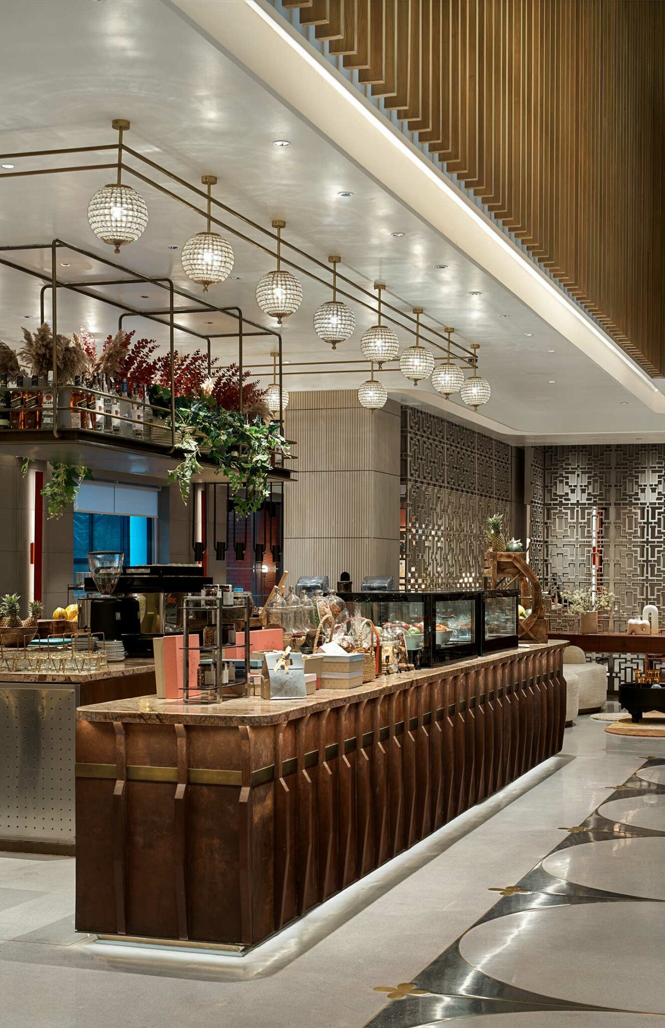 A modern cafe features a long wooden counter with glass display cases, globe pendant lights, and decorative plants, creating a warm, inviting atmosphere.