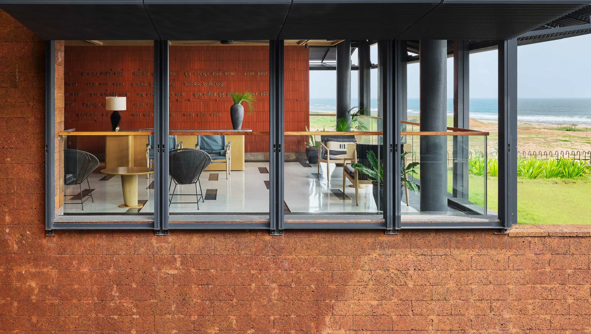 Modern room with large windows overlooking a coastal view. Interior features elegant furniture, plants, and a warm brick wall, conveying relaxation and openness.
