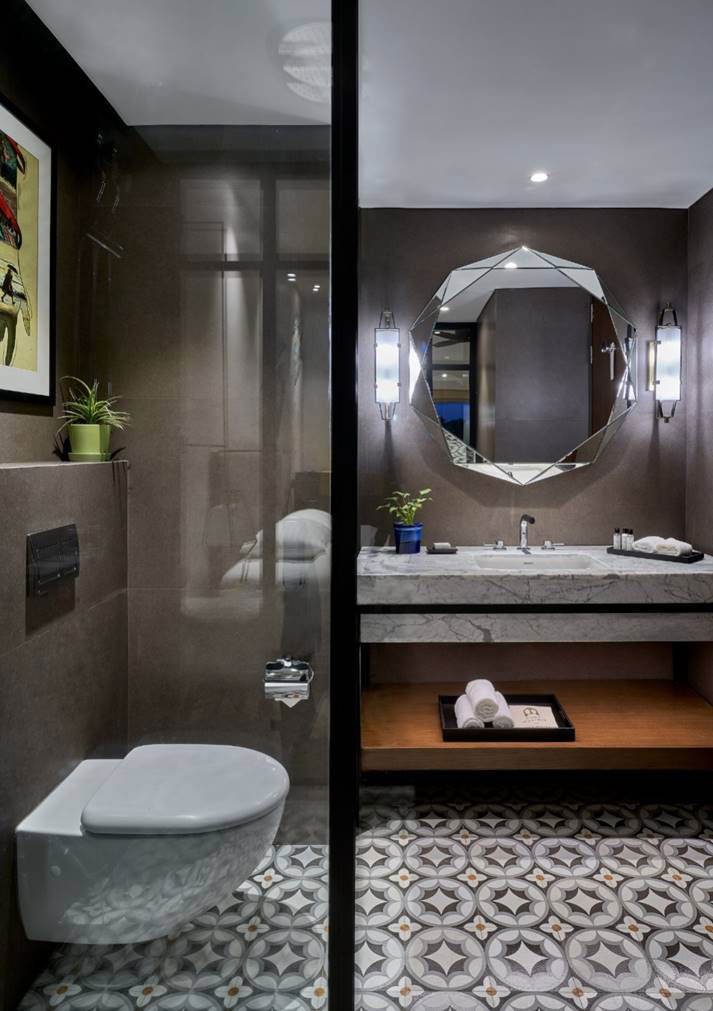 Modern washroom with geometric floor tiles, wall-mounted toilet, large octagonal mirror, plants, and bright lighting, creating a sleek, inviting atmosphere.
