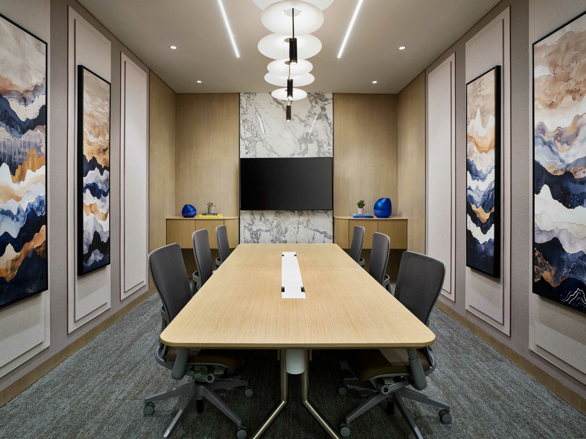 Modern conference room with a long wooden table, Six chairs, abstract wall art, a marble accent wall, and a mounted flat-screen TV. Bright, professional ambiance.
