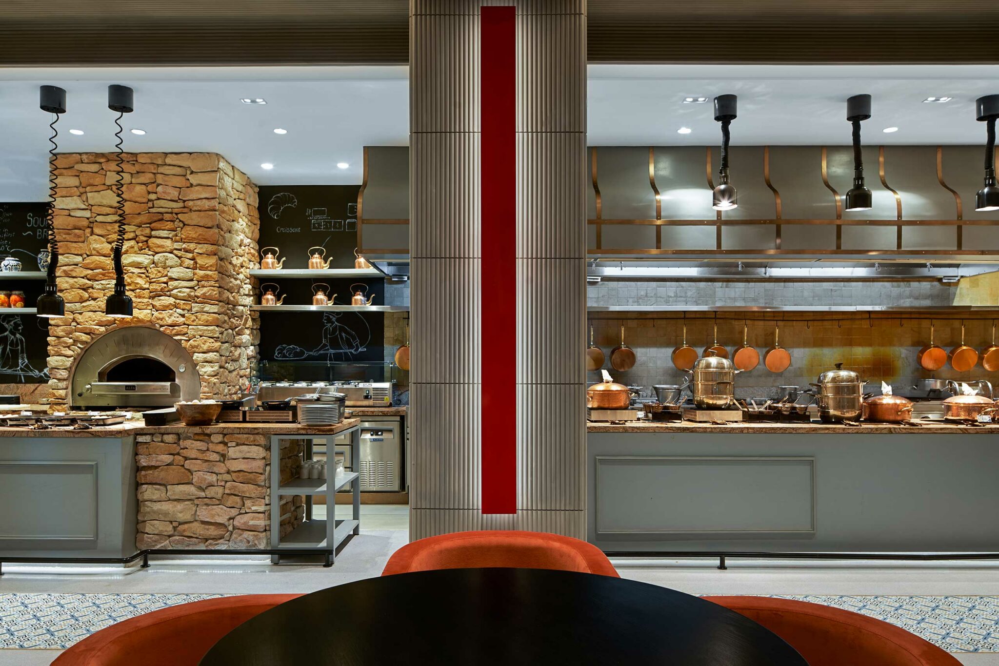 Modern restaurant kitchen with stone pizza oven on the left, hanging lights, copper pots, and pans. Central partition with red accent. Warm, inviting atmosphere.
