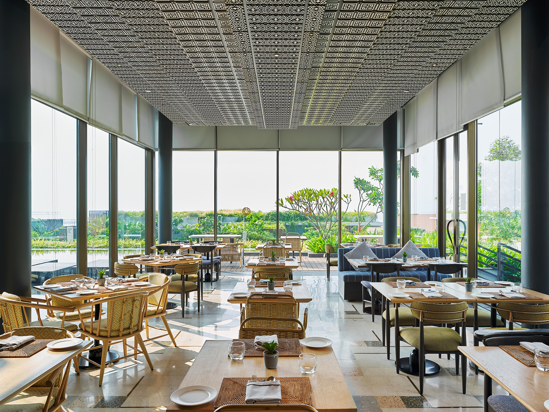 Spacious, modern restaurant with large windows showcasing a lush garden view. Wooden tables, chairs, and soft lighting create a serene dining ambiance.