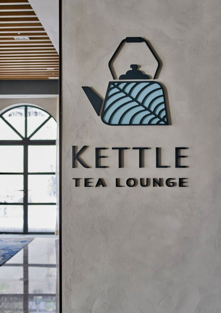 A modern sign for 'Kettle Tea Lounge' on a textured wall, featuring a stylized teapot with a curved line pattern. Bright and inviting atmosphere.