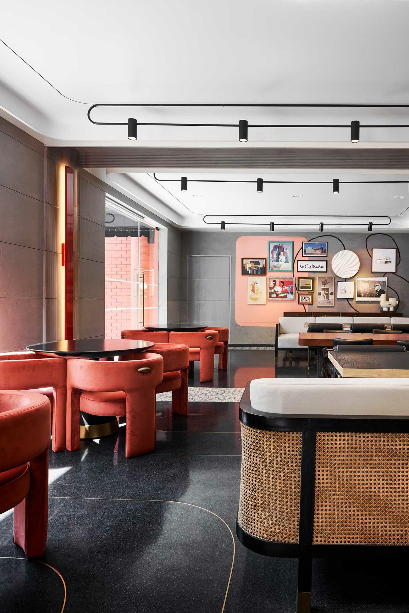 A modern cafe interior with sleek black floors and red velvet chairs around black tables. The walls feature artwork in varied frames, creating a lively atmosphere.
