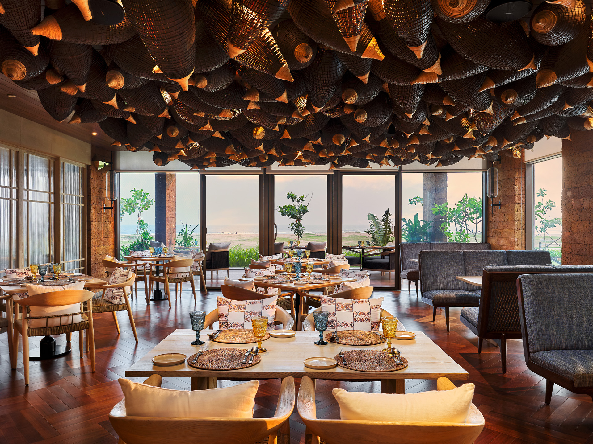 Elegant restaurant interior with a warm, sophisticated vibe. Features woven decor on the ceiling, wooden floors, modern tables, cushioned chairs, and large windows with a scenic view.