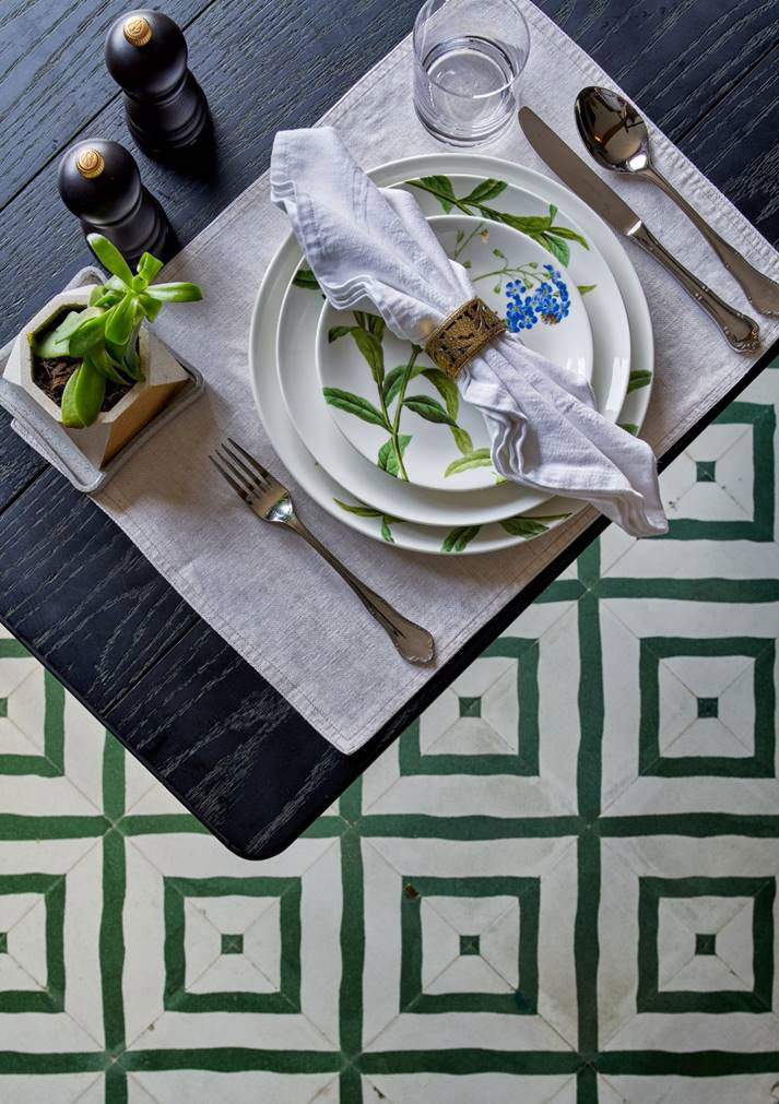 Elegant table setting with leafy plates, folded white napkin, and metallic holder. Nearby, a small potted plant, salt, and pepper shakers sit on a dark table. Green geometric tiles add a bold, stylish touch.