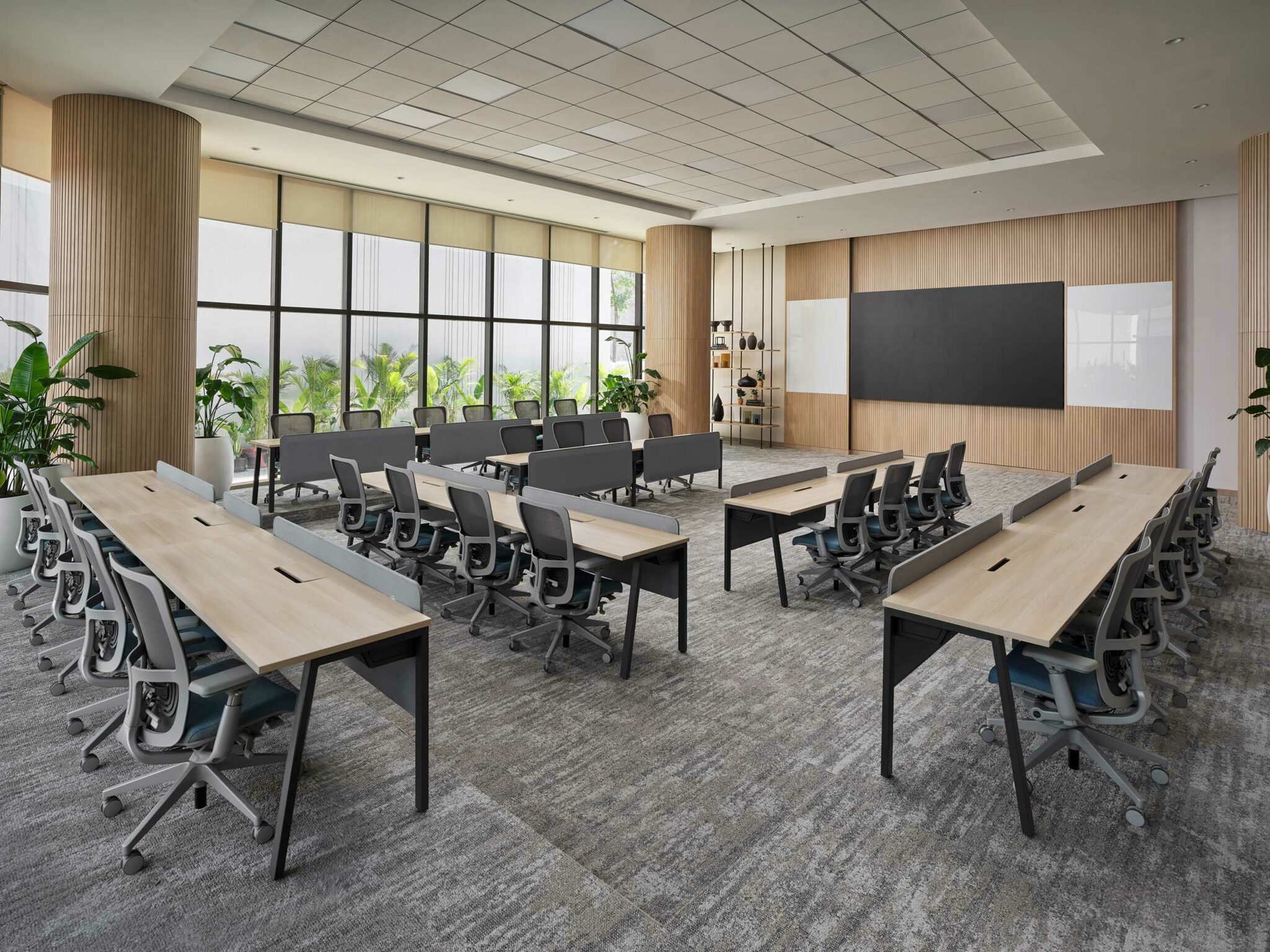 Modern conference room with two rows of wooden desks and gray chairs facing a large blackboard. Tall windows with plants allow natural light, creating a calm ambiance.