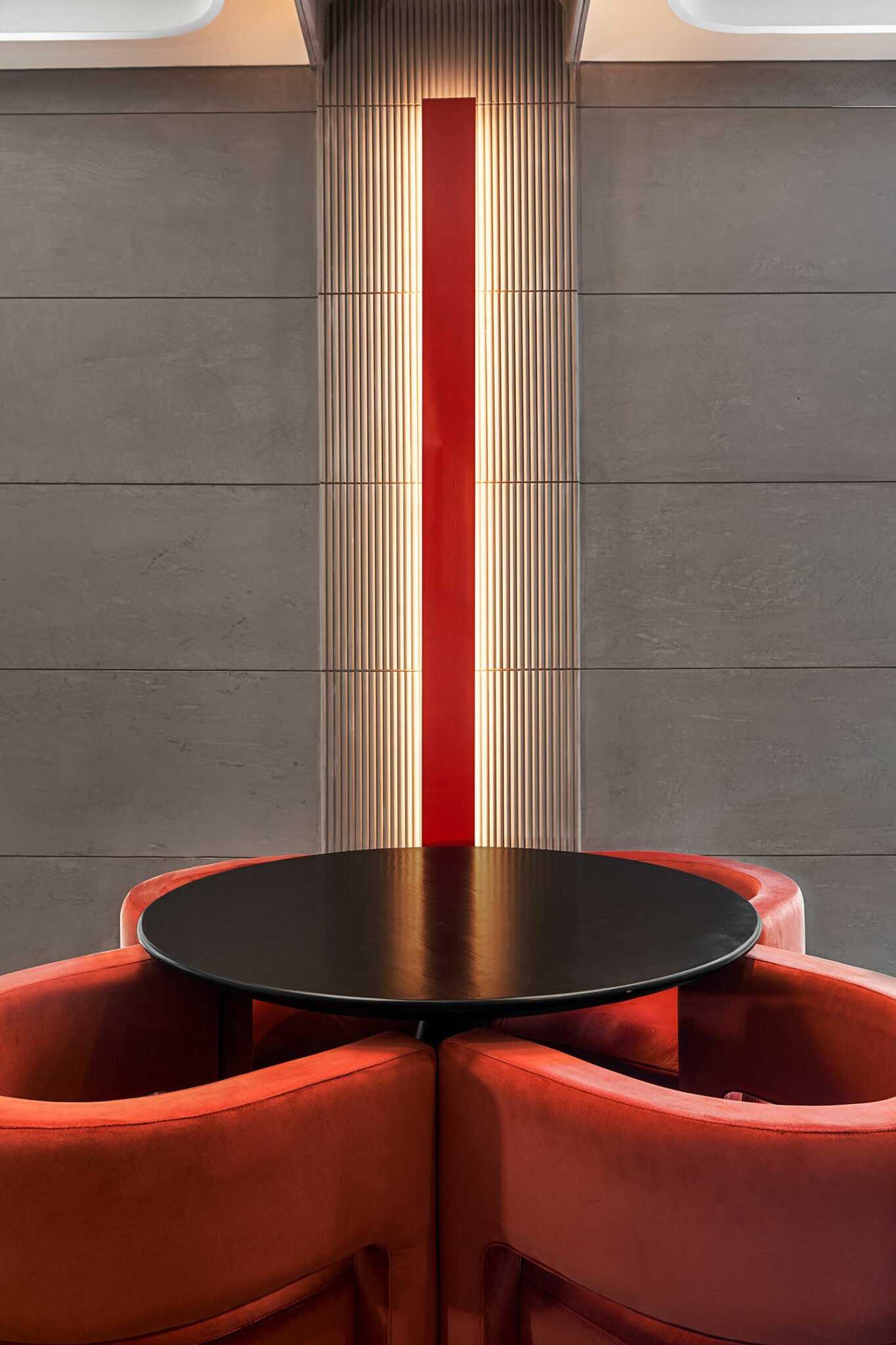 A modern, minimalist setting featuring a round black table surrounded by plush red chairs. A vertical red and illuminated strip accentuates the gray wall.