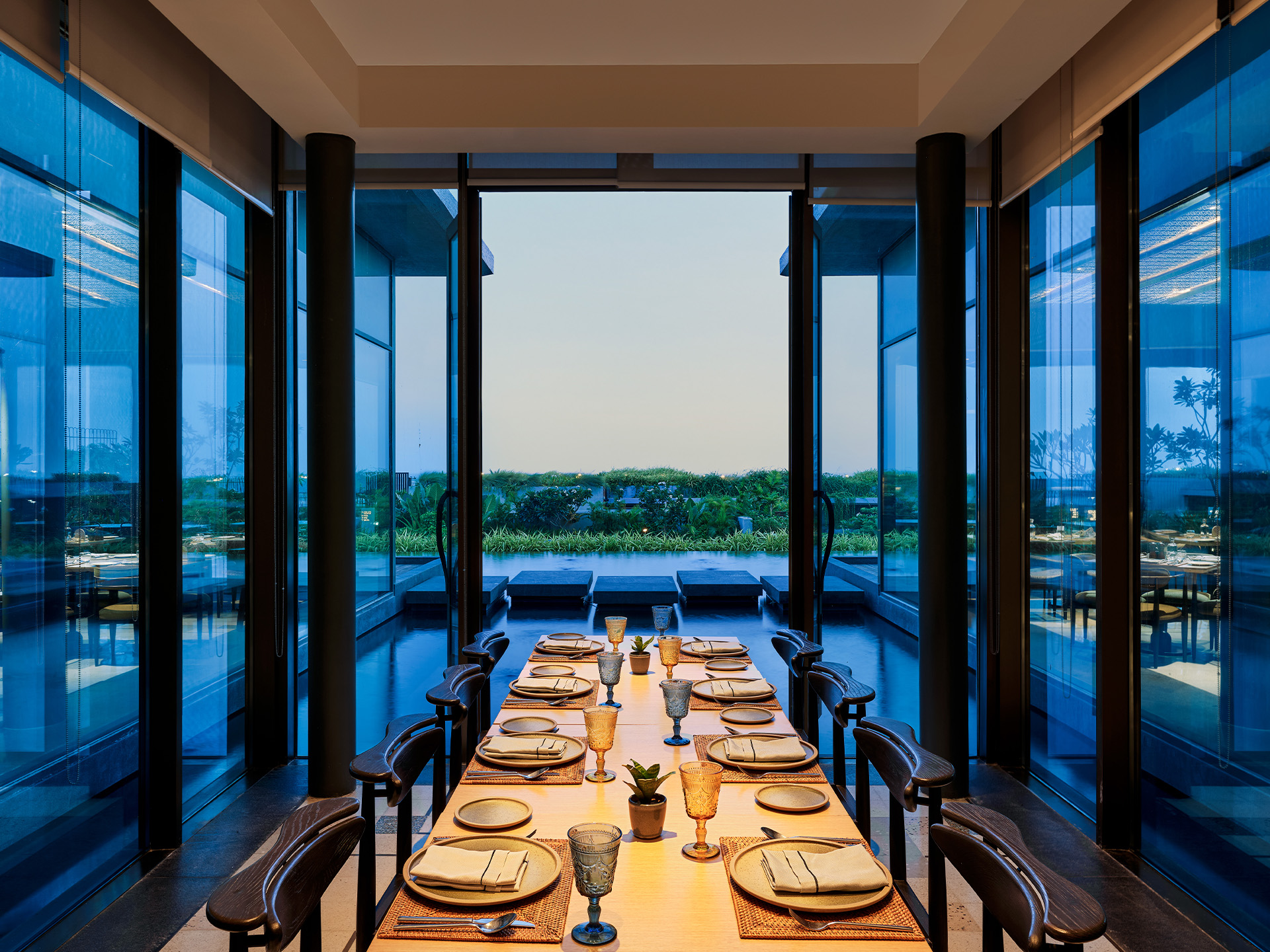 Elegant dining room with a long wooden table set for eight, surrounded by reflective glass walls. A serene outdoor view is visible at dusk.