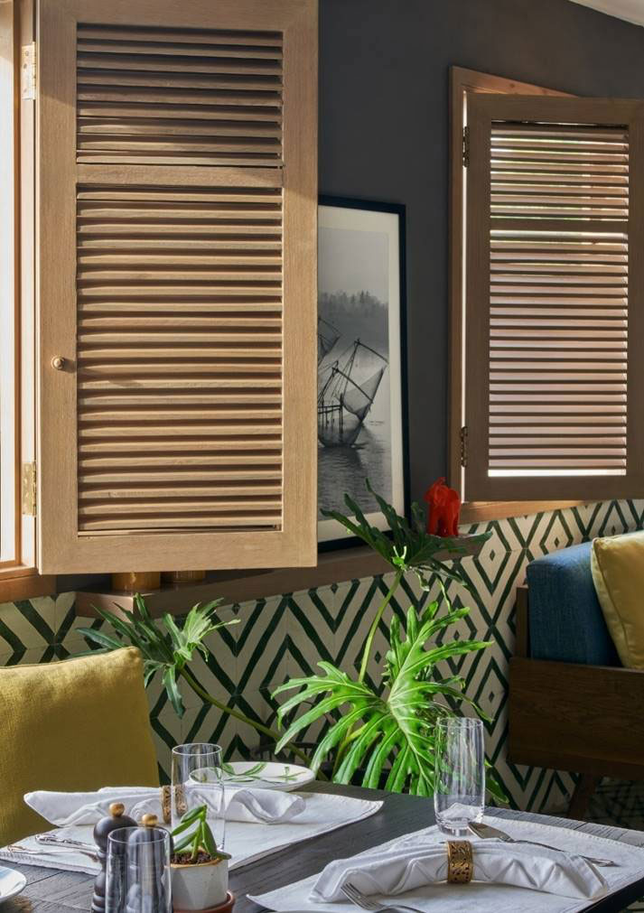 A cozy Tea lounge area features wooden shutter windows allowing sunlight in. A geometric-patterned wall, lush plants, and elegant table setting add warmth.
