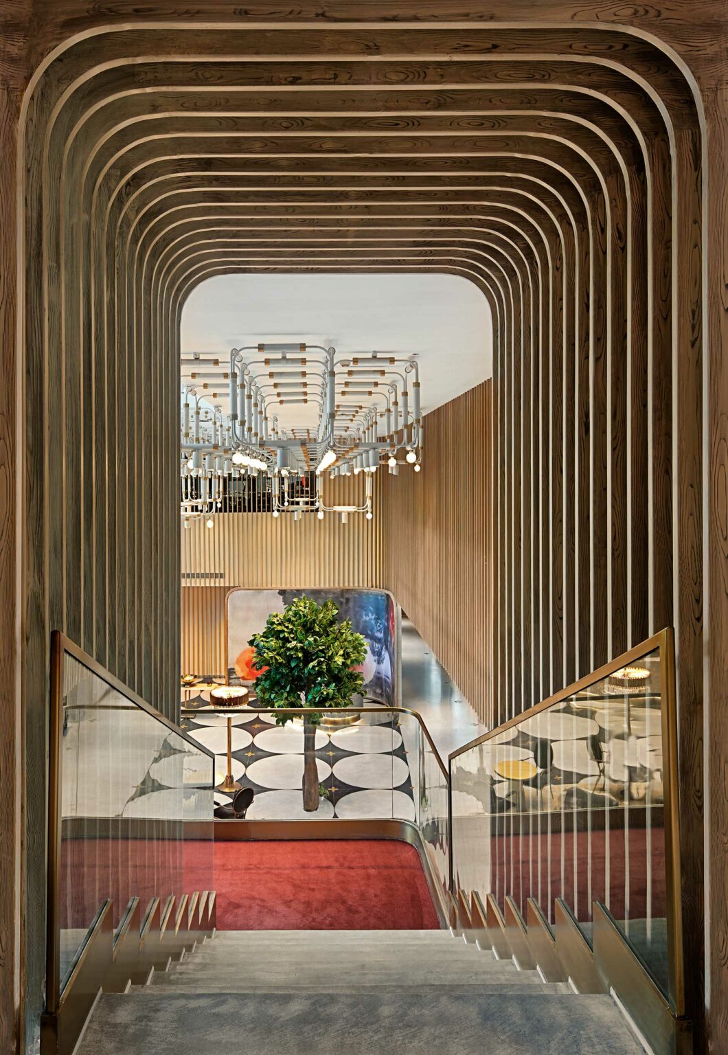 A modern staircase with wooden slats frames a view into a spacious room, featuring geometric floor tiles, a central green plant, and an elegant chandelier.