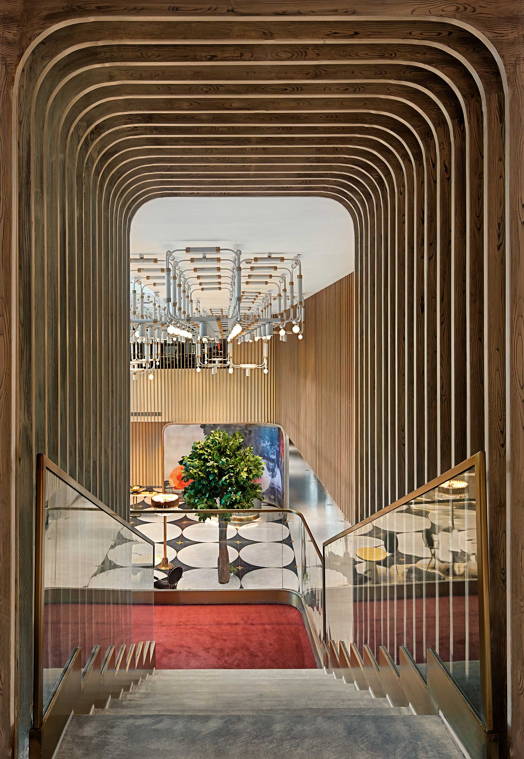A modern staircase with wooden slats frames a view into a spacious room, featuring geometric floor tiles, a central green plant, and an elegant chandelier.