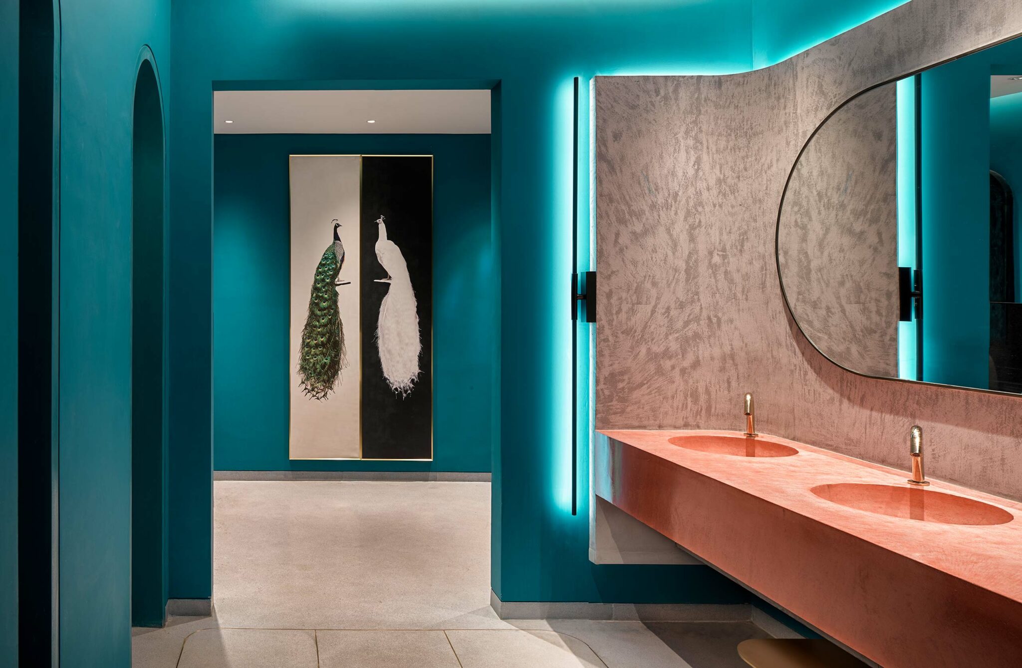 Modern restroom with teal walls, a long pink sink, and circular mirror. Two vertical peacock artworks in the background add elegance.
