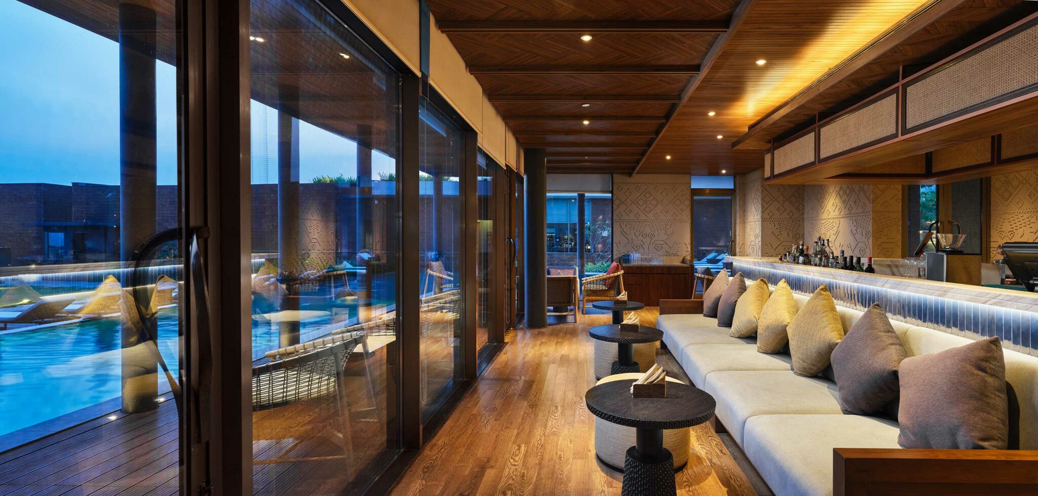 Luxurious lounge with plush cushions, warm wooden decor, and ambient lighting. Glass walls reveal a serene poolside view at dusk, evoking relaxation.