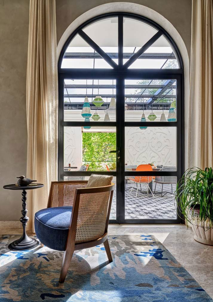 A cozy room with an arch window and beige curtains overlooks a patio. A blue upholstered chair and a round table sit on a patterned rug, with a potted plant nearby.