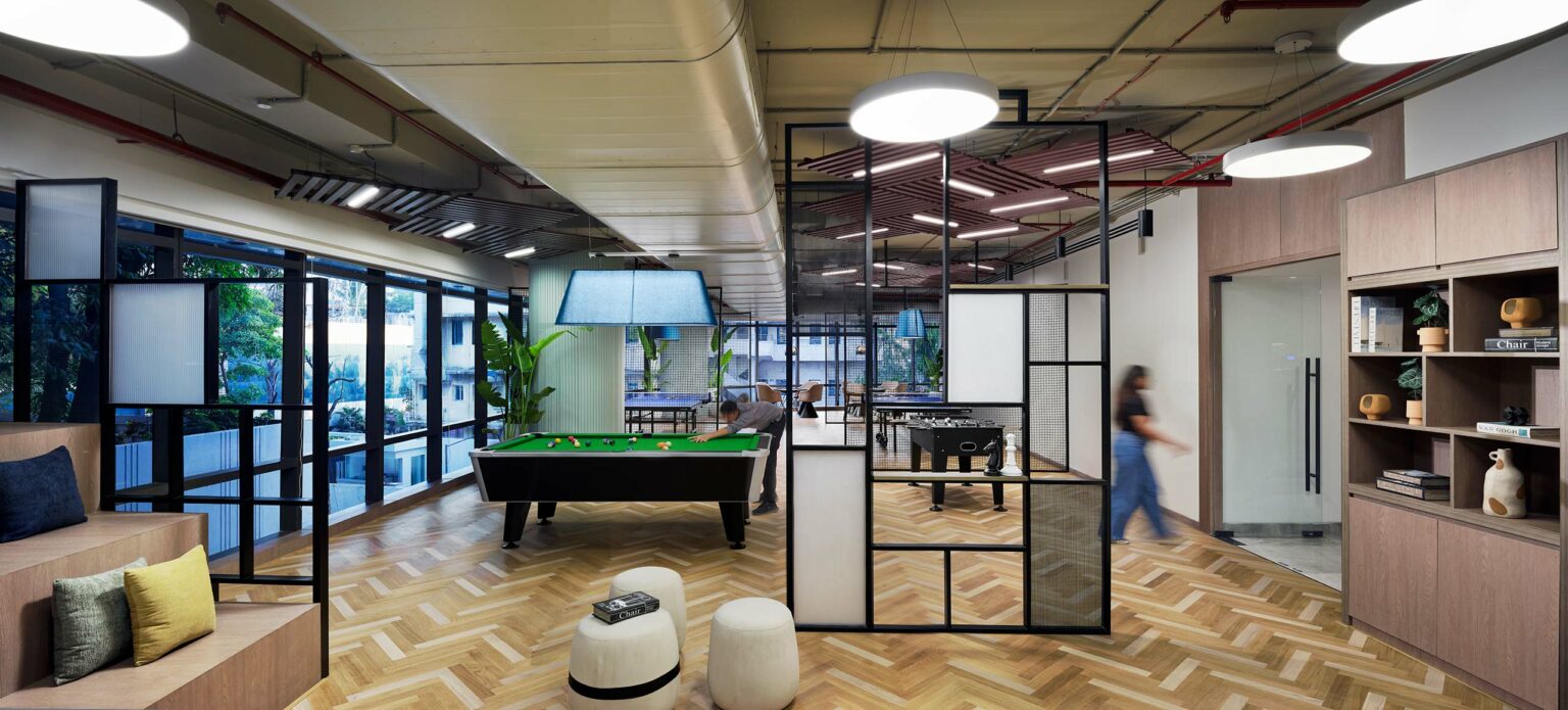 Modern office recreation space with wood flooring, a pool table, and seating area. Large windows provide natural light; a person walks by energetically.