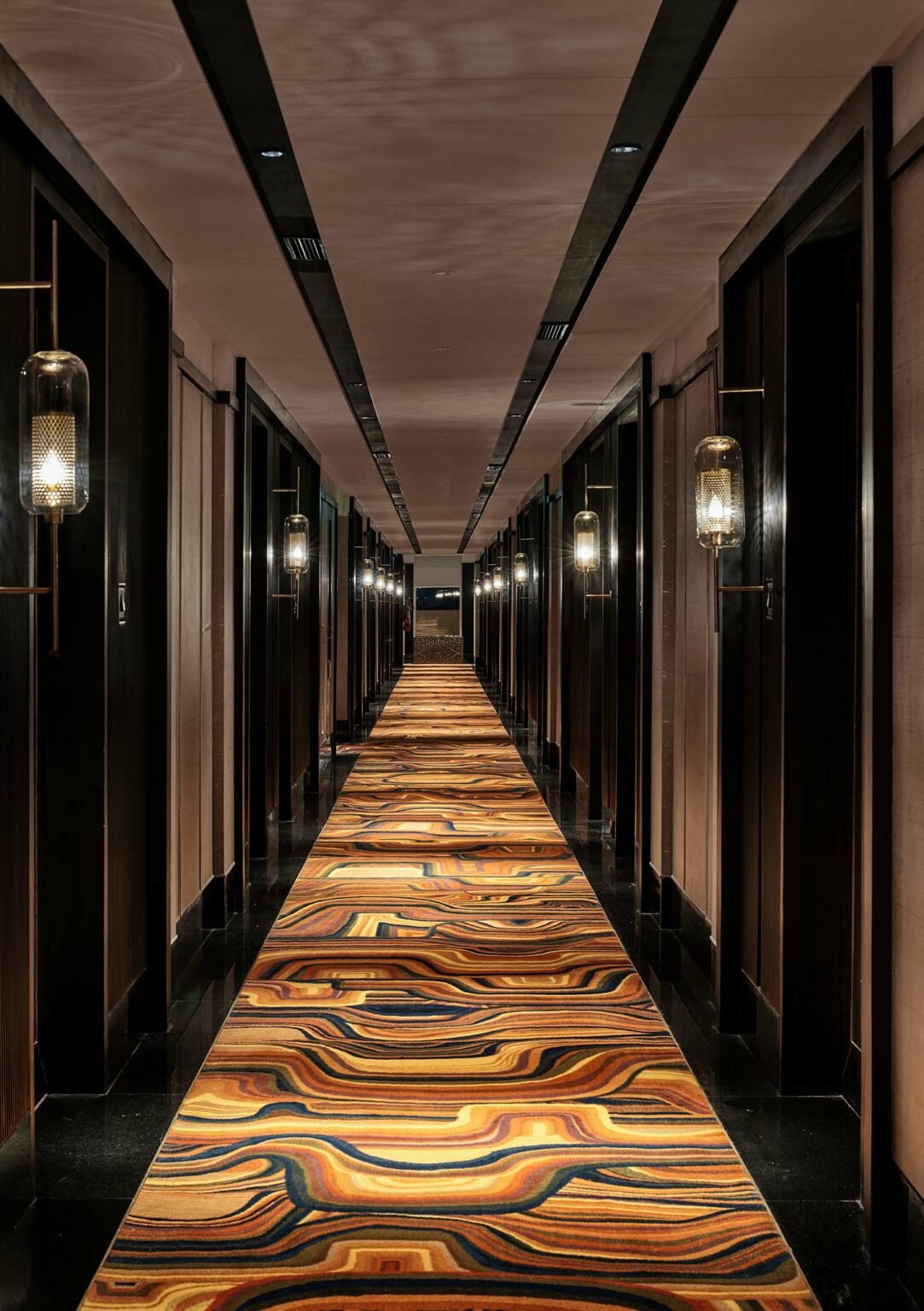 Dimly lit hotel corridor with dark wooden doors on both sides. A vibrant, multicolored carpet with swirling patterns runs down the center, creating a warm, inviting atmosphere.