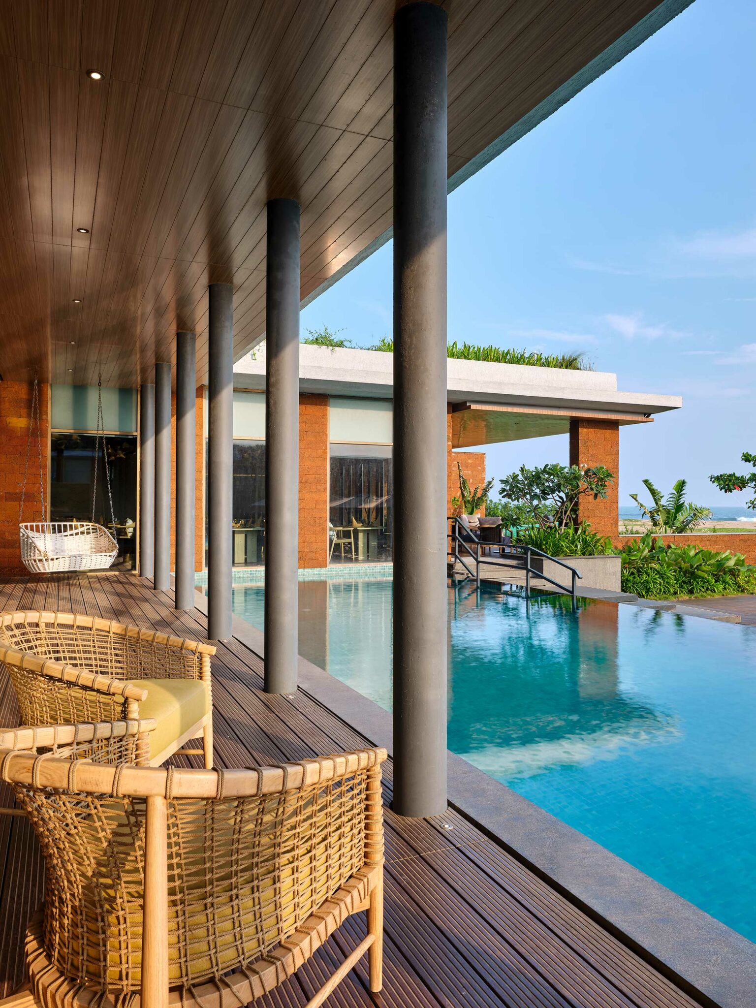 Luxurious poolside view with wicker chairs on a wooden deck, surrounded by modern architecture. Bright, serene atmosphere with clear blue skies.