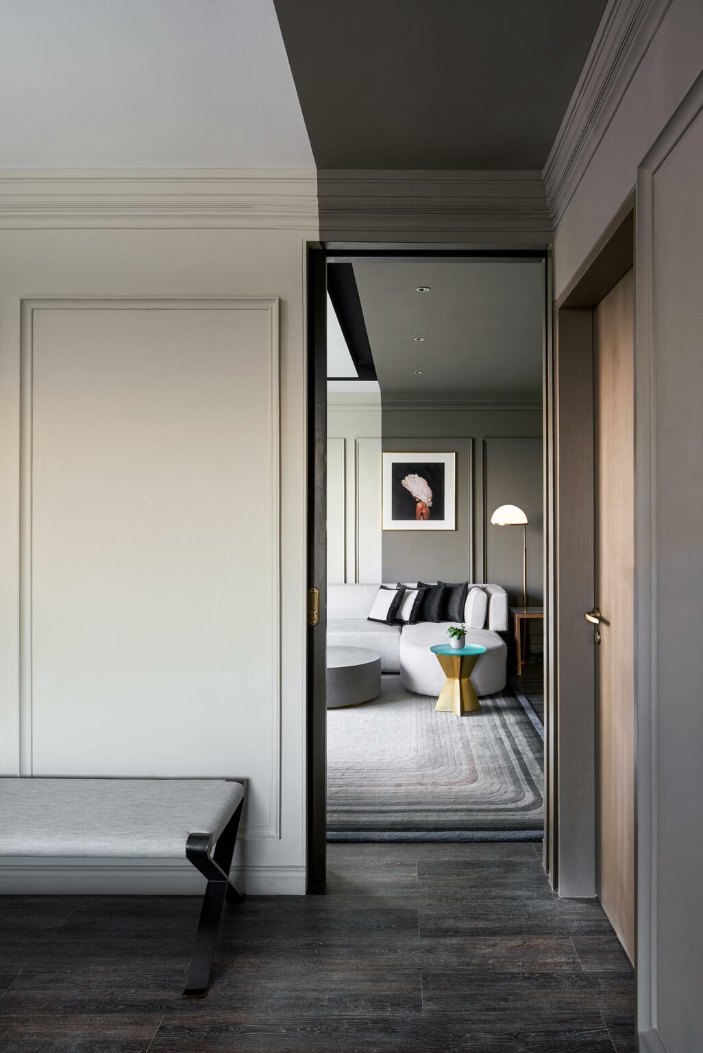 A modern living room seen through an open doorway, featuring a gray sofa, gold side table, floor lamp, and monochrome artwork. The space feels calm and elegant.