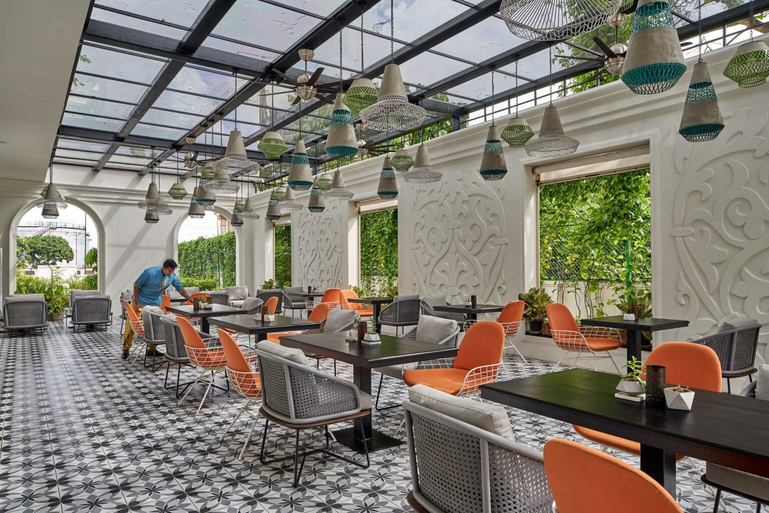 Stylish outdoor Dining with a glass roof and ornate white walls. Orange and gray chairs surround dark tables on geometric patterned floors. A person tidies a table, creating a welcoming ambiance.