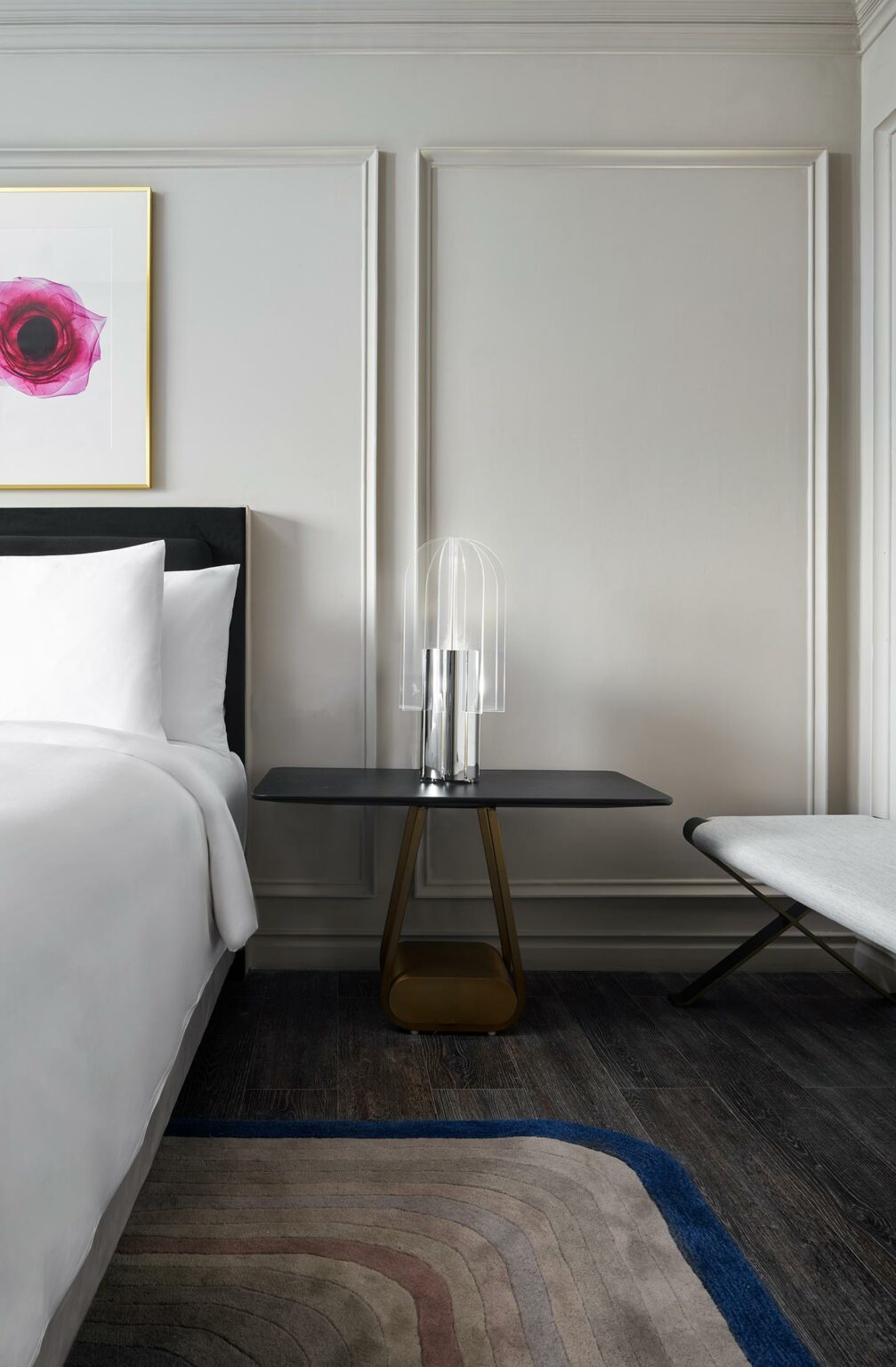 Minimalist bedroom scene with a white bed, black side table, and modern lamp. Abstract pink art and muted tones create a serene atmosphere.