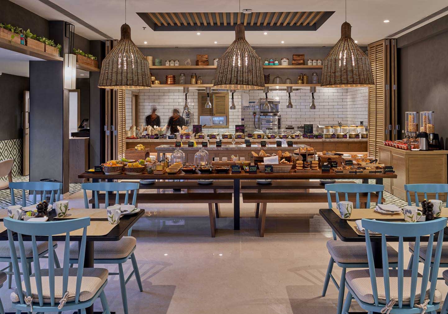 Modern breakfast buffet with wooden tables and blue chairs. Abundant pastries, fruits, and cereals under warm pendant lights. Inviting and cozy ambiance.