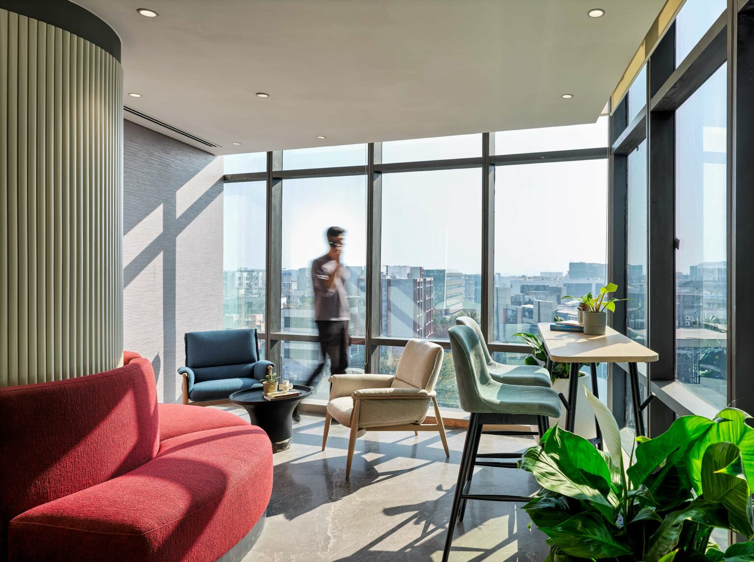 Sunny office lounge with modern furniture, including a red couch, blue and beige chairs, and green plants. A man, walking by large windows.