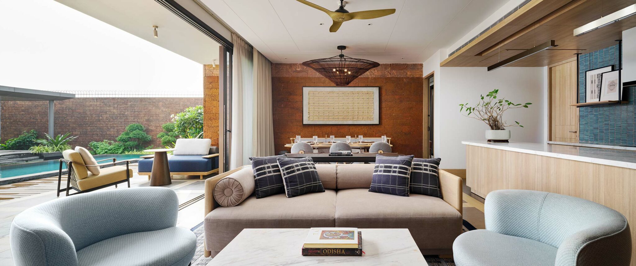 A modern living room with a luxurious feel; features a beige couch, light blue chairs, a marble coffee table, and a pool view through wide-open doors.