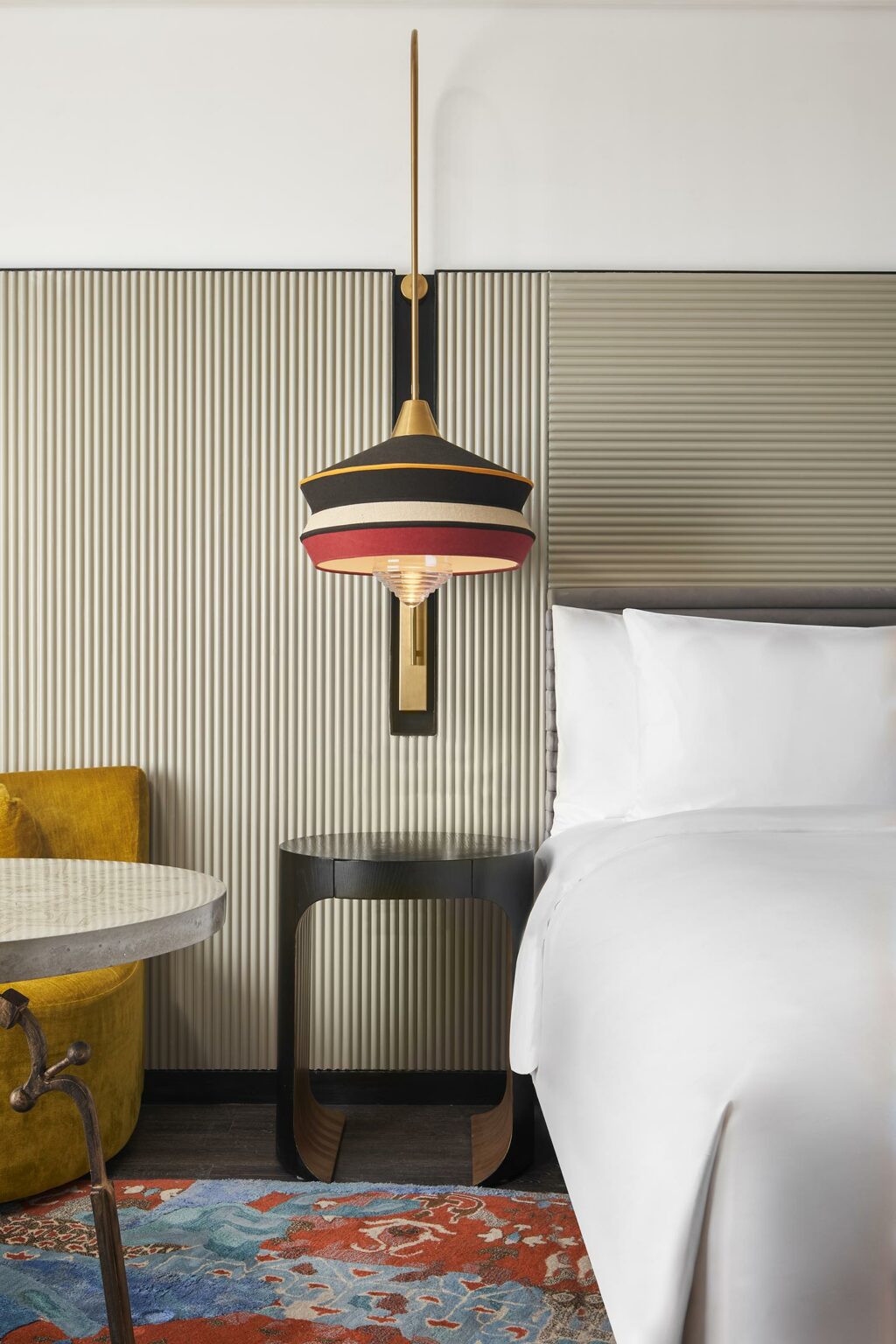 Modern hotel room featuring a white-sheeted bed, red and black hanging lamp, mustard yellow chair, abstract rug, and a sleek black side table.