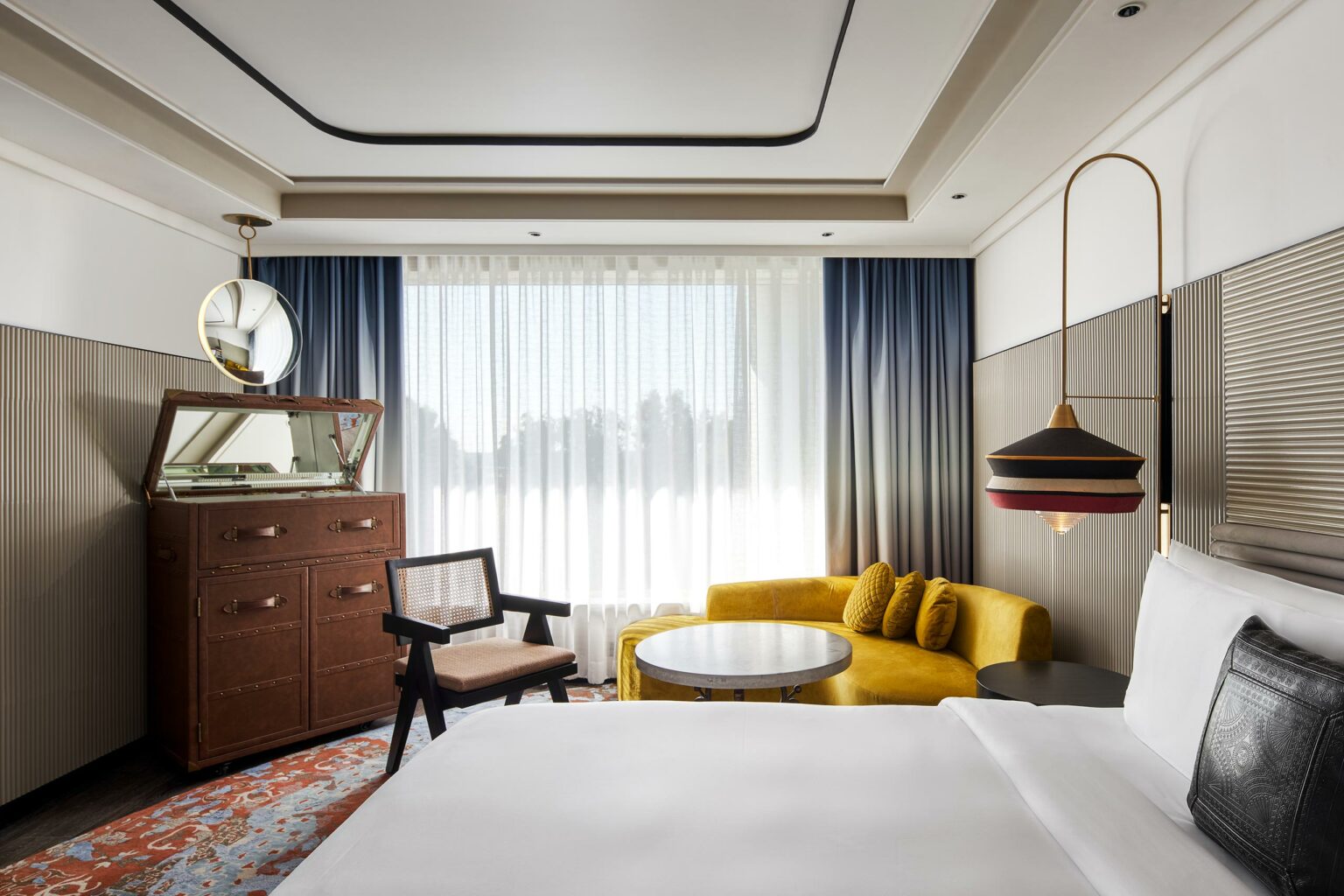 Elegant hotel room with a modern design, featuring a yellow sofa, round table, and chic lighting. A large window allows natural light to illuminate the space.