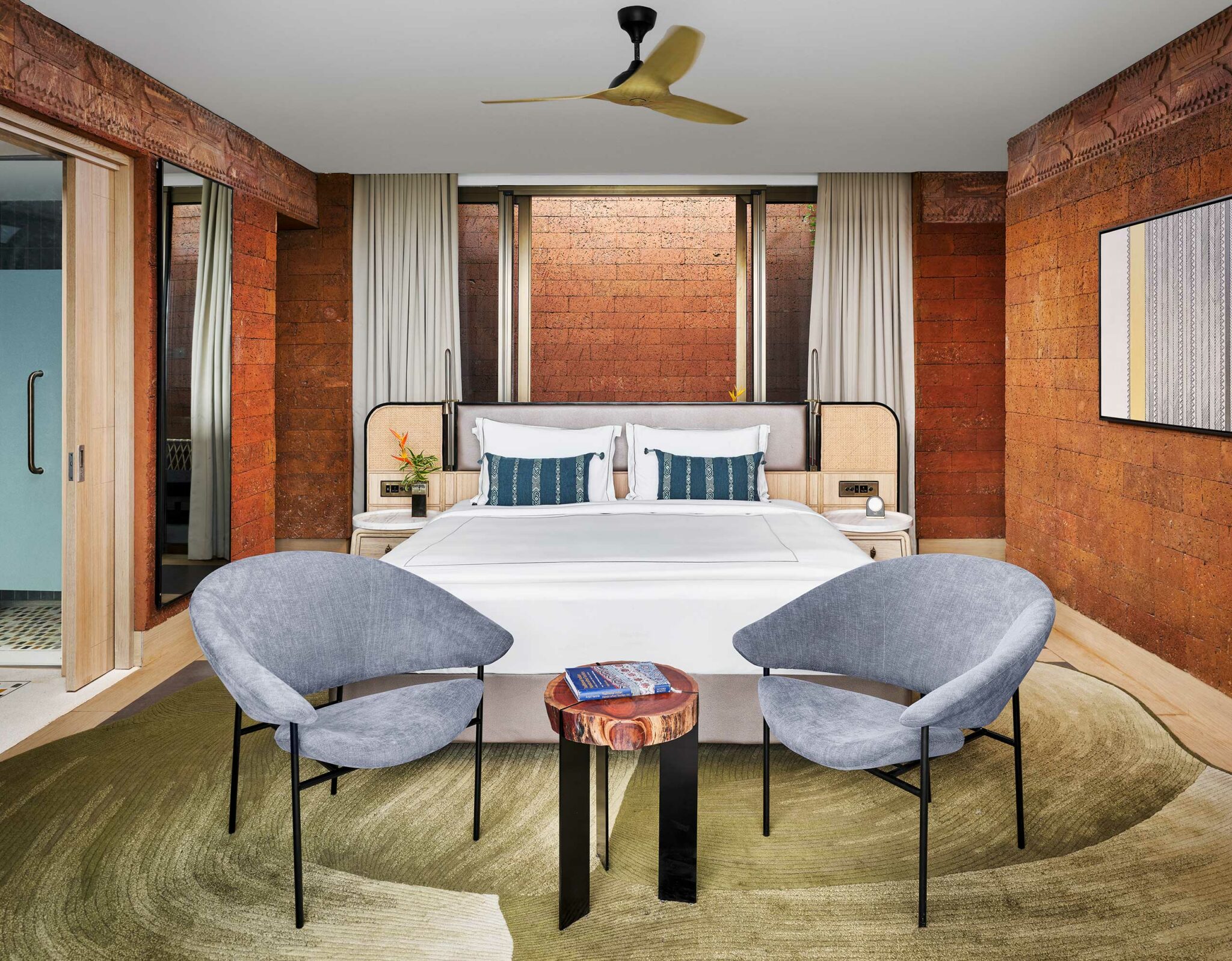 Modern hotel room with a white bed, two blue cushions, and light wood furnishings against a rustic brick wall. Two gray chairs and a small wooden table on a green rug add elegance.