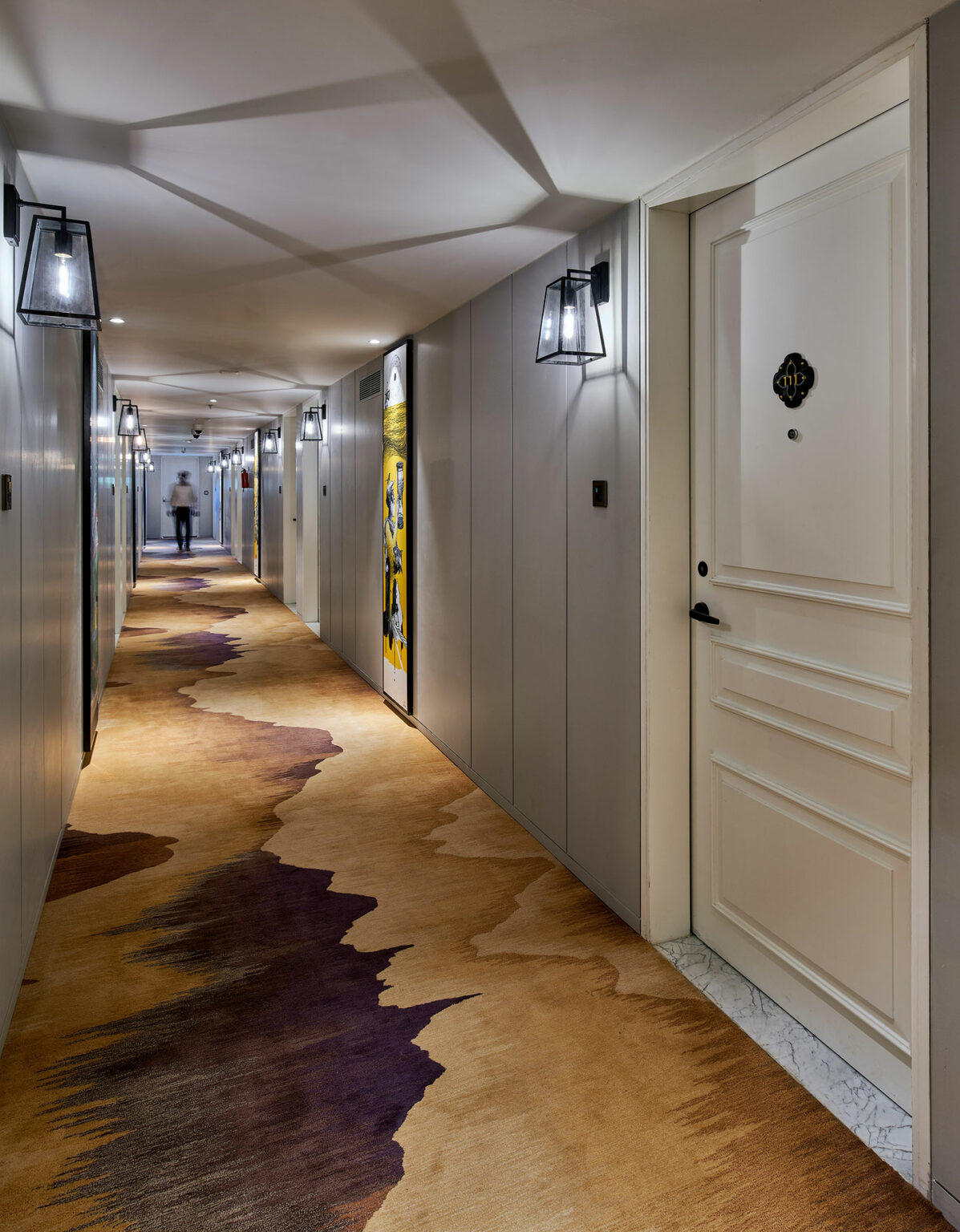Modern hotel corridor with warm lighting, featuring patterned carpet in earthy tones. White doors line the walls, with a person walking in the distance.