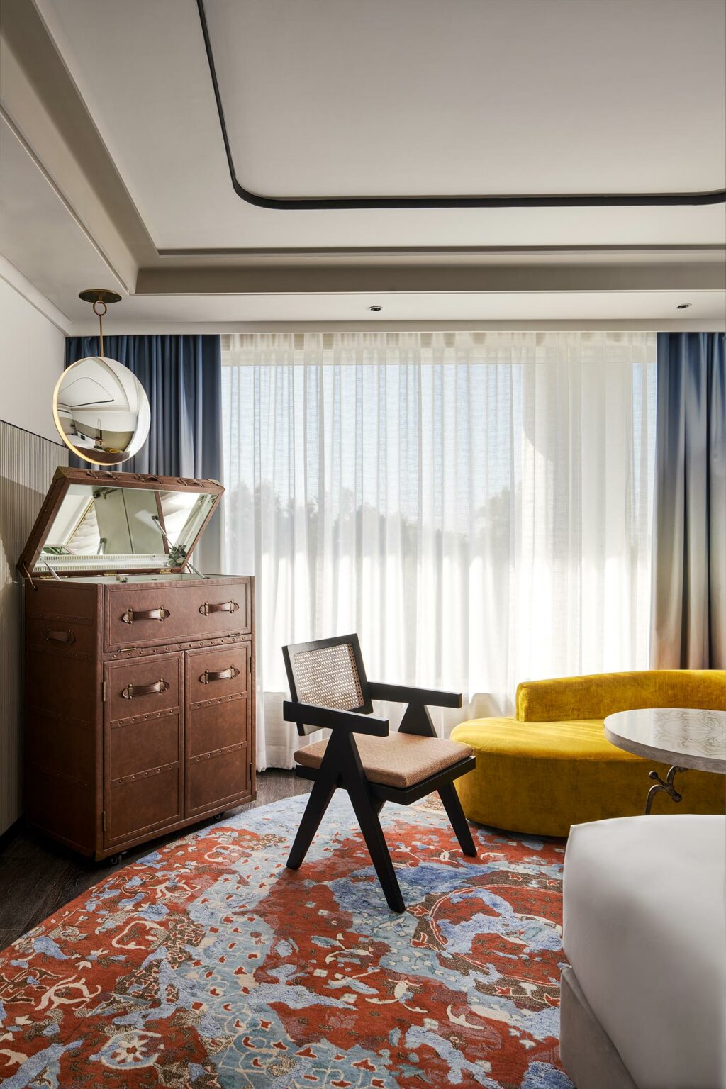 A cozy hotel room with a rustic brown dresser, a round mirror, a yellow sofa, a black chair, and a colorful red and blue patterned rug. Soft light enters through sheer curtains.