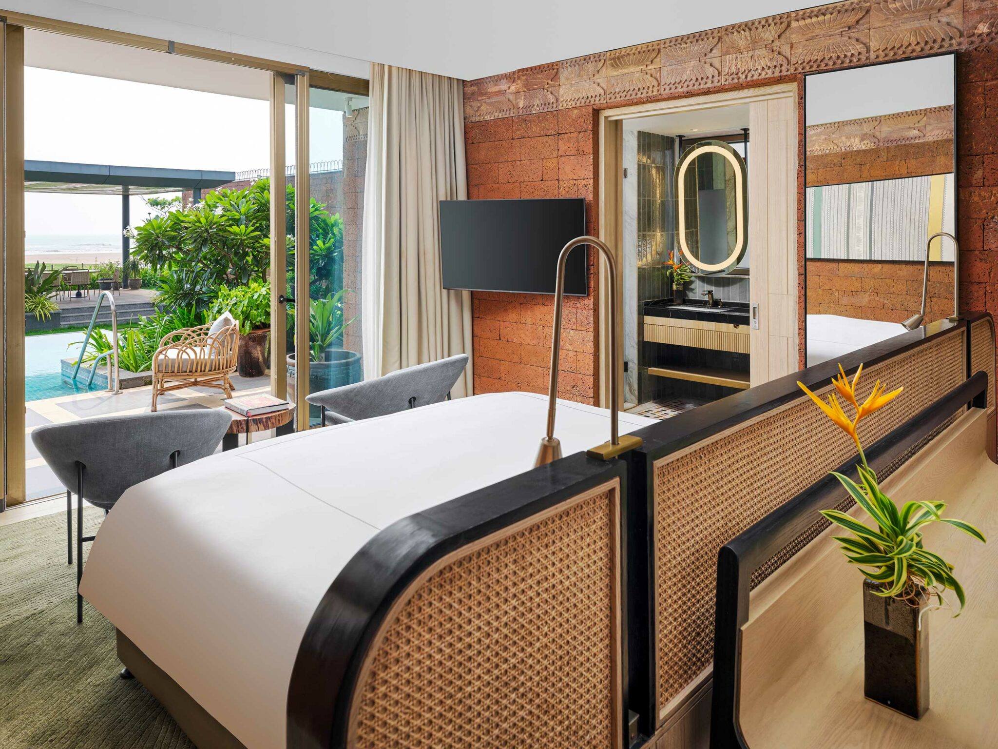 Modern hotel room with a pool view. Features a large bed, woven headboard, sleek decor, plants, and a bathroom with an illuminated mirror, evoking luxury.