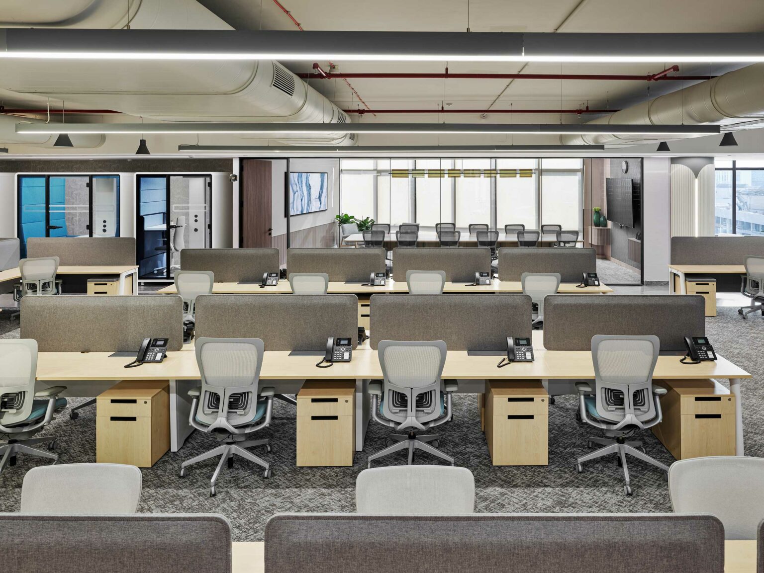 Spacious, modern office with rows of desks and gray chairs. Telephones on each desk. Bright lighting and large windows create an open, airy atmosphere.