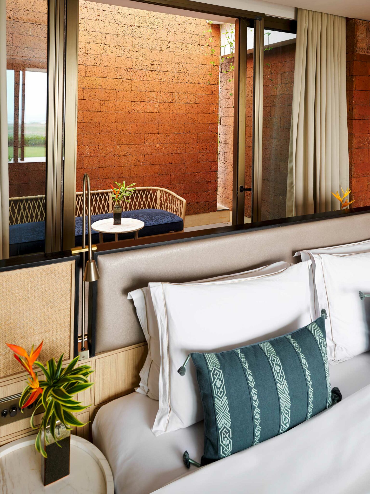 Hotel room with a large bed featuring white sheets and a blue patterned pillow. A glass wall reveals a cozy patio with a wicker chair and small table.