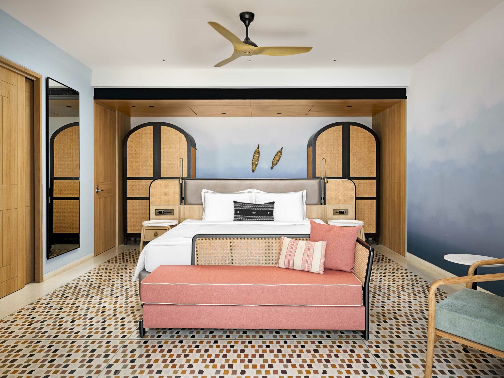 Stylish hotel room with a modern double bed, pink sofa, mosaic floor, and a ceiling fan. Natural tones and soft lighting create a calm ambiance.