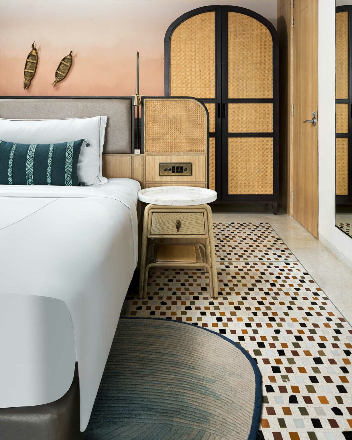 Modern bedroom with a stylish white bed, teal cushion, rattan wardrobe, and woven wall decor. Warm tones, geometric rug, and cozy ambiance.