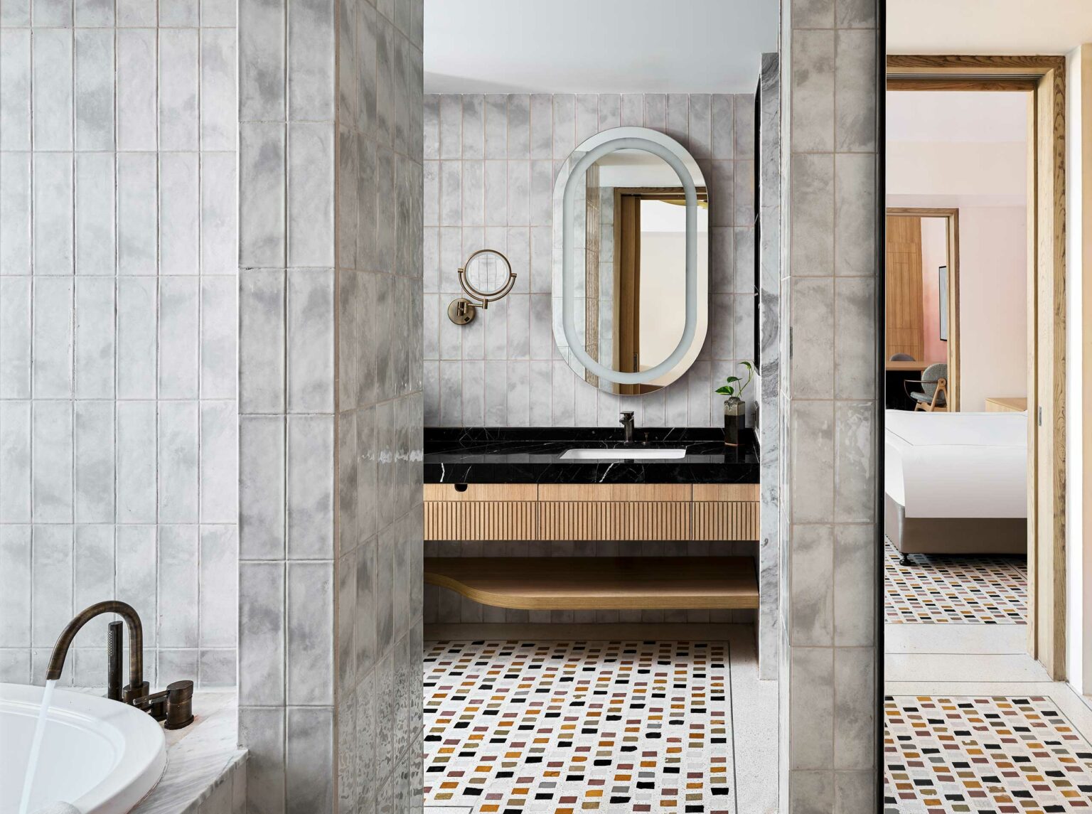 Modern bathroom with gray tiled walls and mosaic floor. Features a black countertop with an oval mirror, round magnifying mirror, and visible adjoining bedroom. Clean and elegant design.