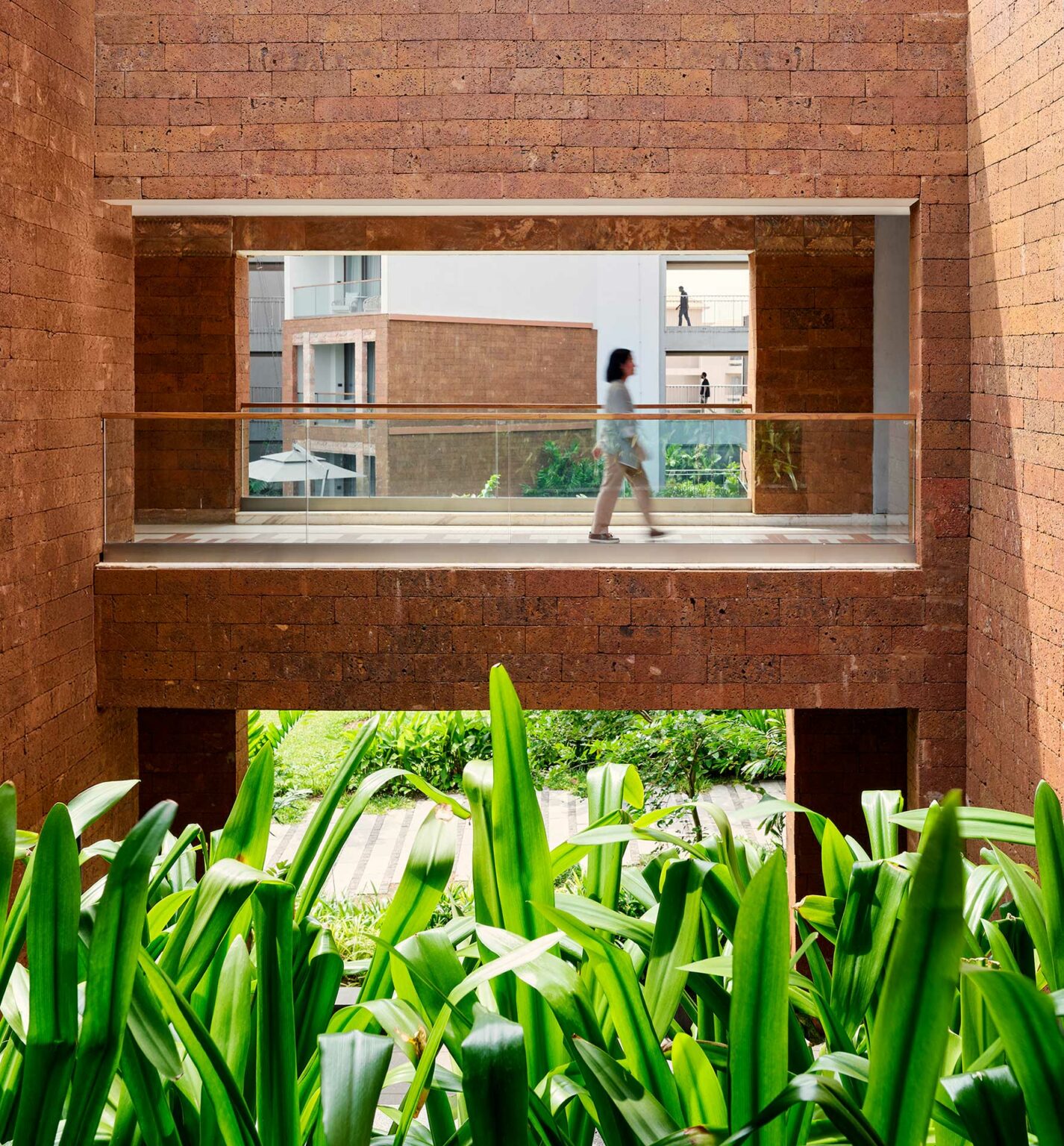 A person walks in a modern building's glass corridor framed by red brick walls. Green plants in the foreground create a fresh, serene atmosphere.