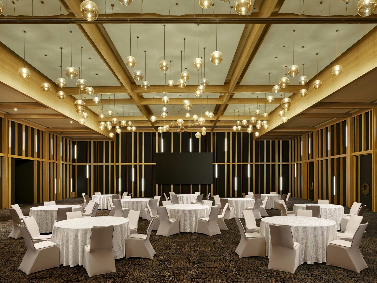 Elegant banquet hall with round tables draped in white tablecloths and cushioned chairs. Warm lighting from globe chandeliers creates a sophisticated ambiance.