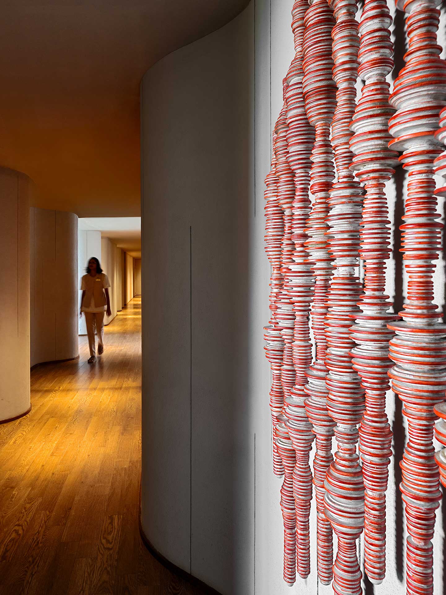 Curved hallway with wooden floor, featuring a wall with an artistic installation of stacked red and white discs in the foreground; a person walks in the background.